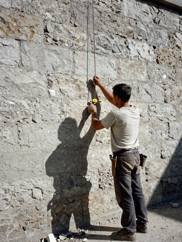 A professional mason using a plumb line against a raw stone wall, harsh daylight casting sharp shadows, gritty documentary style, 35mm