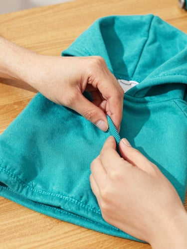 Close-up of hands carefully sewing a vibrant teal pet hoodie on a wooden table, warm golden light, focus on fabric texture and precise stitching, cozy workshop environment