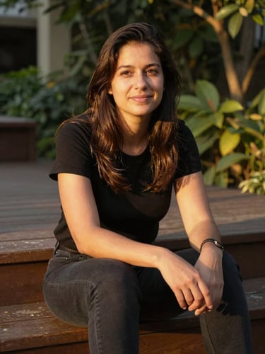 Close environmental portrait, photographer seated on sun-warmed wooden steps in a garden, looking slightly off-camera with a relaxed half-smile, late afternoon golden light falling across her face and hands, soft amber bokeh of foliage behind her, documentary framing