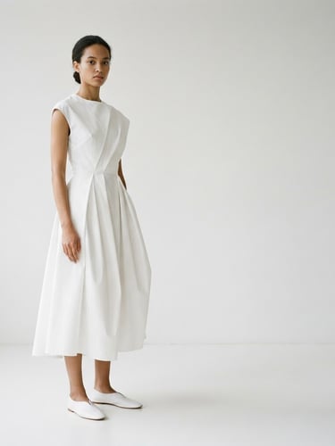 A minimalist fashion photograph of a model wearing a structurally precise, unembellished white garment, soft natural daylight, clean white background, focus on silhouette and drape, plenty of negative space to the right.