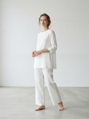 Model wearing a minimalist white cotton-silk tunic with clean lines, standing in a spacious, light-filled studio with soft diffused natural daylight, plenty of negative space, 85mm lens, sharp focus.