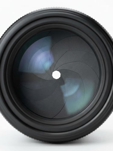 Extreme close-up of a professional camera lens front element, glass reflections, dust-free mechanical iris, high-key studio lighting, white backdrop