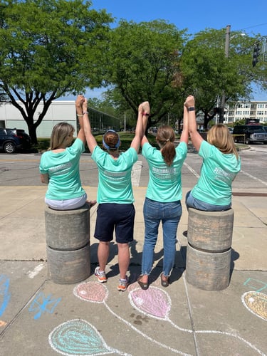Four people in teal Peacemakers Needed shirts holding hands and raising arms on a sidewalk with chalk art.