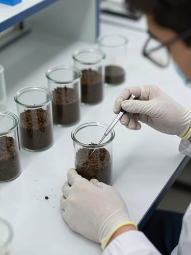 Close-up overhead view of a soil science laboratory in Herat: glass vials containing soil samples on a white bench, a technician's gloved hands holding a measuring instrument, bright studio lighting, documentary framing emphasizing process and precision