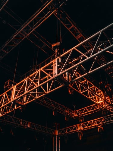 Close-up of a complex metal truss structure under dramatic stage lighting, deep shadows, vibrant orange light accents, architectural lines, wide-angle, sharp details, 35mm