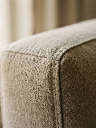 Macro close-up of tight, flawless fabric stitching on a Renovo sofa, soft natural window light casting gentle shadows, highlighting the tailored upholstery texture, warm stone neutrals.