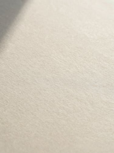 A wide, quiet macro photograph of printed black ink on textured cream paper, soft overcast daylight casting low-contrast shadows, showing the tactile grain of the paper.
