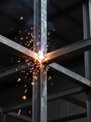 Close-up of structural welding on heavy metal framing, bright orange sparks flying, dark industrial background, dramatic shadows.