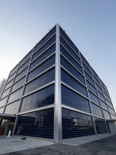 Low-angle architectural perspective of a modern steel-and-glass logistics terminal in Dubai, crisp daylight, silver highlights, deep navy shadows.