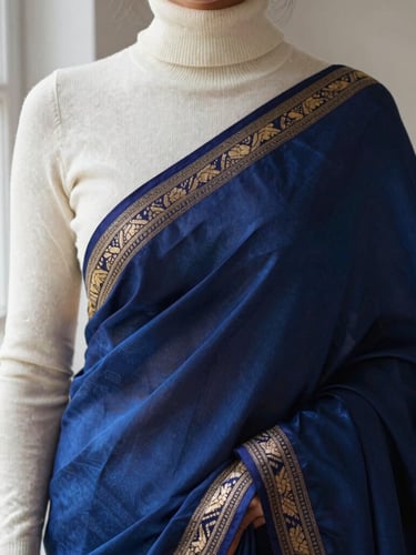 Close-up editorial shot of a deep blue silk saree layered under a cream cashmere turtleneck sweater, soft window light, focusing on the rich gold embroidery details.