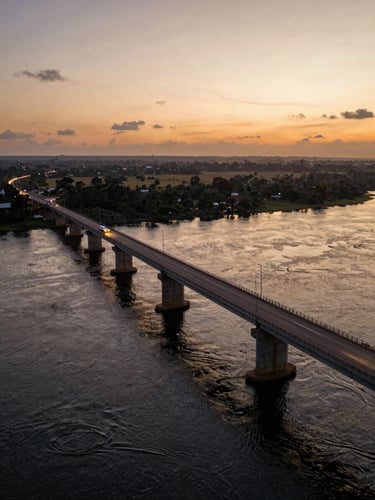 Low-angle cinematic drone shot of a completed bridge over a wide Central African river at dusk, concrete piers rising from dark water, warm gold-orange sky behind the structure, headlights of vehicles crossing visible as light streaks, vast riparian landscape stretching to the horizon