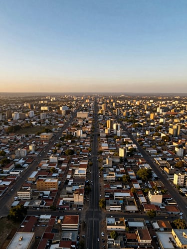 Wide panoramic aerial drone shot looking straight down over a Cameroonian city grid at golden hour — road networks and building footprints rendered as precise geometric patterns, the terrain glowing amber and deep shadow, a single surveying tripod silhouette visible at the far left edge, vast sky above the right third