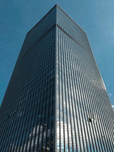 Low-angle architectural photograph of a modern glass skyscraper corner piercing a vibrant blue sky, crisp daylight, sharp reflections of clouds on glass.