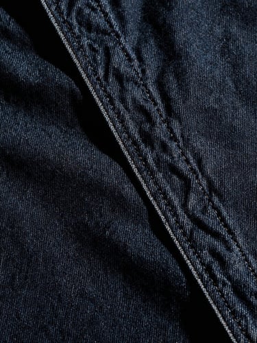 Extreme close-up of heavy raw selvedge denim seams, dark indigo fabric texture, high-contrast directional studio light, deep shadows