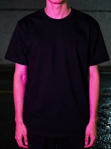 Low-angle cinematic shot of a model wearing a black graphic tee under neon pink rim lighting, wet pavement, dark concrete background, 35mm film grain