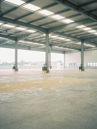 Wide-angle architectural film photography of a vast, empty, modern logistics warehouse with high ceilings and automated systems, soft natural overcast light, desaturated cool tones with warm sand accents, 35mm wide-angle, no people.