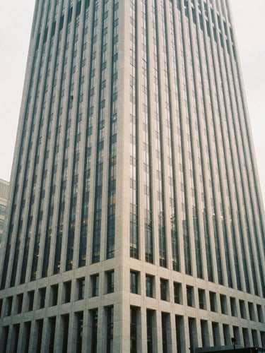 Wide-angle architectural film photography of a modern, high-rise corporate building in a bustling city financial district, soft natural overcast light, desaturated cool tones with warm sand accents, 35mm wide-angle, no people.
