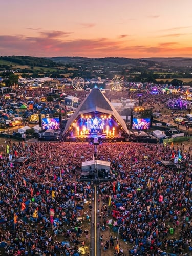 Aerial wide-angle drone shot of a massive outdoor music festival at sunset, vibrant stage lighting, expansive crowd, high-production setup, professional 35mm lens