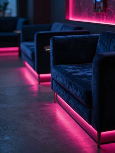 cinematic editorial photography, dramatic low-key lighting, deep navy and neon fuchsia accents, high contrast, 35mm. Close-up detail of a luxury hotel lobby lounge at dusk, velvet seating, glowing neon accent lighting casting fuchsia reflections on polished concrete floors, empty space, atmospheric depth.