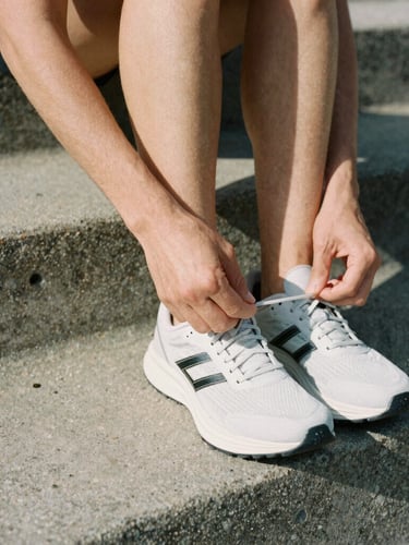 Tight, high-contrast crop of an active professional tying minimalist running shoes on a raw concrete step, natural morning light, medium-format film aesthetic.
