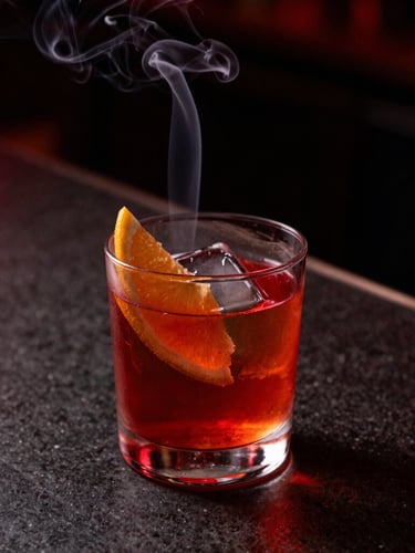 Close-up of a smoking Negroni cocktail on a dark stone counter, dramatic red neon highlight, high contrast, 35mm lens