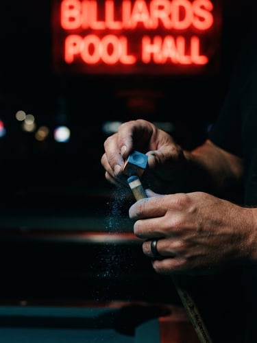 Moody low-key close-up of hands chalking a pool cue under a sharp red neon light, deep shadows, high-contrast editorial style