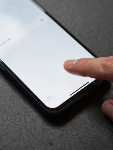Extreme close-up, top-down framing — a single thumb pressing the display of a matte-black Android phone against a dark graphite desk surface, screen mid-transition showing a crisp UI, even fluorescent work-light casting no shadows, tight crop on the interaction moment