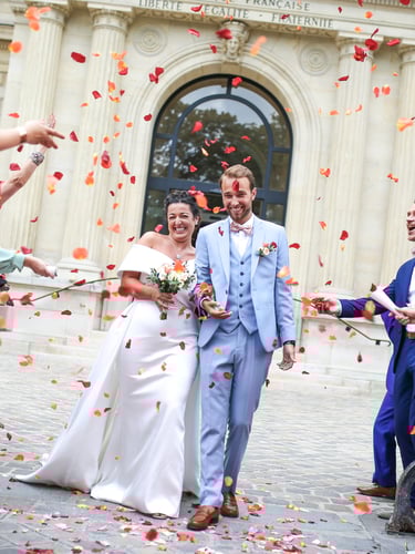 Smiling bride and groom exit a French building while guests toss orange rose petal confetti.