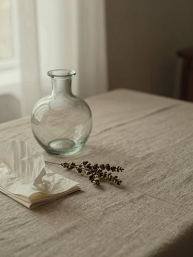 Close-up flat-lay on warm linen cloth, a single hand-blown glass vessel beside folded tissue paper and a sprig of dried botanicals, diffused window light from the left, muted warm tones, quiet negative space to the right