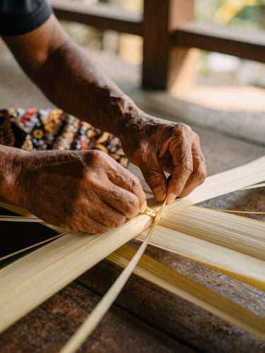 A close-up of local Balinese artisan hands carefully weaving lontar leaves in a sunlit wooden pavilion, warm earthy tones, soft shadows, 35mm lens.