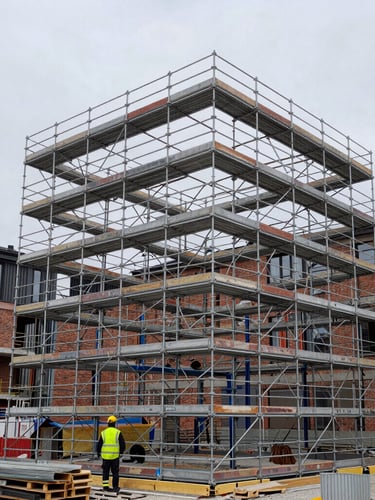 Wide environmental shot of a heavy steel scaffolding structure erected against a modern brick building under overcast Stockholm morning light, workers in high-visibility safety gear visible at the base.
