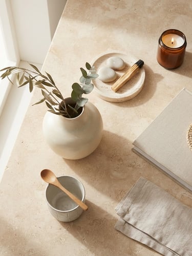 Overhead styled flat-lay of minimalist home decor accessories on a warm stone surface, bright natural window light, soft diffused shadows