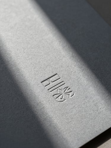 Extreme close-up of embossed stationery on heavy cool ash gray paper stock, hard directional natural light casting a sharp diagonal shadow across the texture.