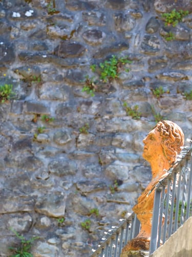 A classical terracotta bust sculpture on a wrought iron railing against an ancient stone wall background.