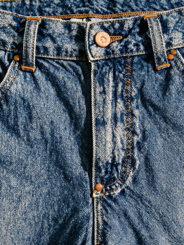 Close-up of heavy raw denim texture showing triple-stitched seams and copper rivets under harsh natural daylight, high contrast, 35mm film photography.