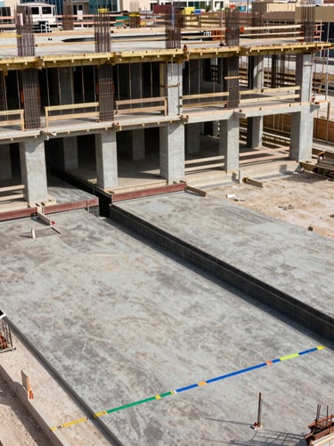 Wide overhead plan-view of an active Dubai construction site showing phased slab pours at different levels, rebar placement visible on upper deck, MEP conduit runs marked with colour tape on lower slab, daylight, minimal correction, no workers in frame