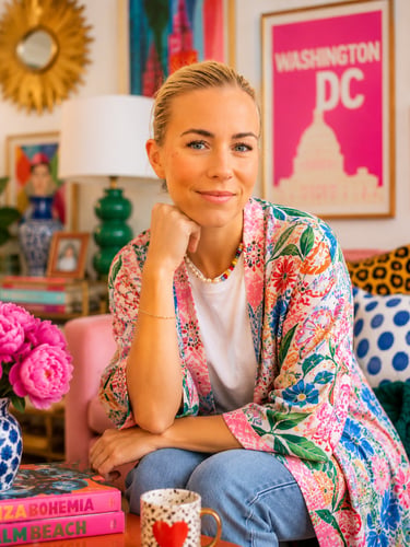 A blonde woman in a floral kimono sits in a colorful living room with vibrant decor and wall art.