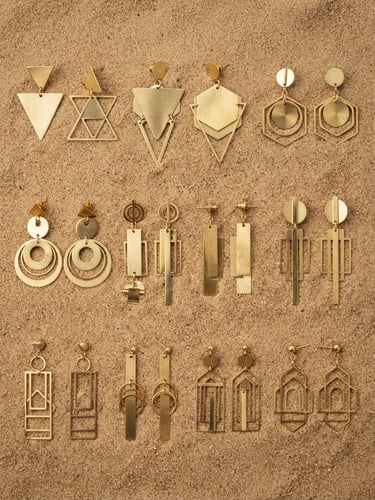 Macro photography of polished gold and brass geometric earrings arranged neatly on a warm sand-textured surface, sharp focus, professional industrial lighting, 35mm