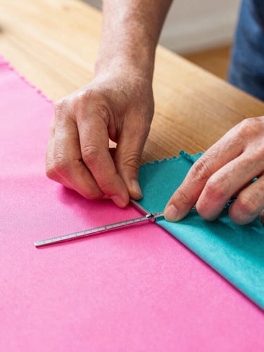 Close-up of artisan hands carefully measuring and cutting vibrant pink and teal fabrics on a wooden table, warm natural light, shallow depth of field.