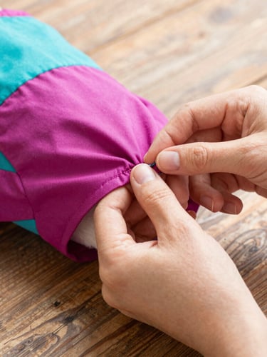 Close-up of hands carefully stitching a colorful magenta pet dress on a rustic wooden table, bright natural light, showing fabric textures.