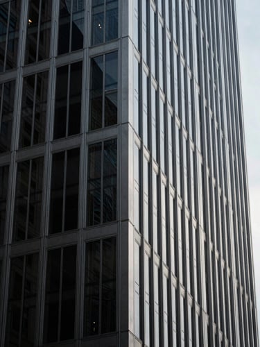 Minimalist architectural close-up of a modern steel and glass skyscraper corner in New York, dramatic high-contrast lighting, deep shadows, clean lines against a pale sky.
