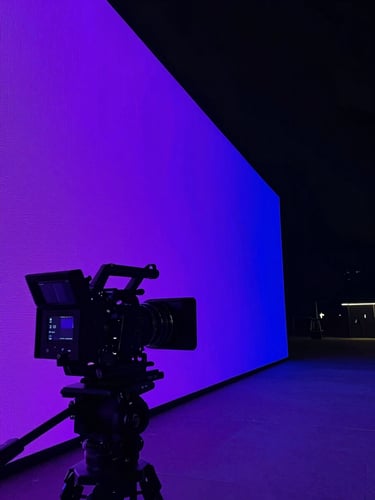 Panoramic cinematic wide shot of a digital campaign launch moment — a large format LED wall displaying a bold brand visual in electric purple and blue, camera rig in foreground silhouetted against glowing screen, low angle, dramatic contrast, studio environment to the left, generous dark negative space on the right third