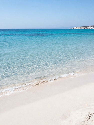 Wide environmental shot of a sun-drenched Greek beach with crystal-clear turquoise waters and white sand, midday sun reflecting on water, bright coastal photography