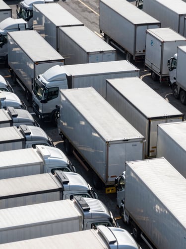Clean geometric overhead shot of an organized fleet yard with aligned commercial transport trucks, overcast daylight, sharp metallic reflections
