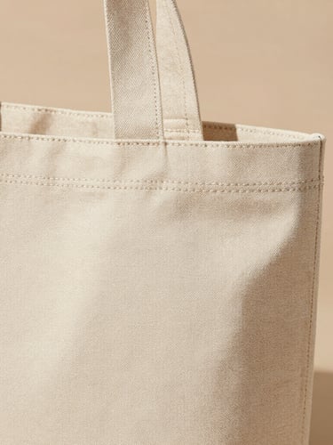 Close-up of premium canvas tote bag stitching, clean minimalist aesthetic, soft natural daylight, warm sand tone background, high-end editorial look, 35mm