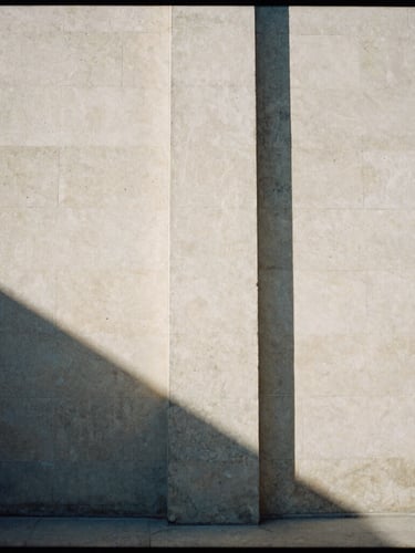 Stark architectural concrete pillar casting a long, sharp shadow on a warm stone wall, high-contrast natural light, minimalist editorial style, 35mm film grain