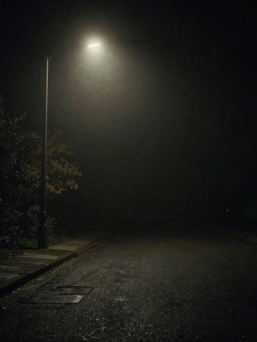 A dark empty street at night under a single flickering streetlight, heavy fog, high-contrast shadows, eerie atmosphere, cinematic 35mm film.