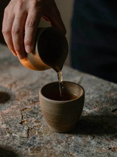 A close-up of hands pouring tea into a raw clay cup on a weathered stone surface, warm candle glow, soft shadows, moody editorial 35mm film