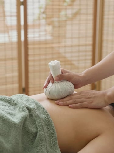 A professional therapist gently placing warm herbal compress on a shoulder, soft natural lighting filtering through bamboo screens, warm editorial photography, 35mm, muted moss green and warm stone tones