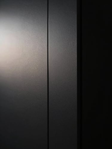 Extreme close-up of a matte black architectural wall panel, razor-thin silver seam running vertically off-center, dramatic chiaroscuro side-lighting from the left casting a deep shadow gradient across the surface, near-total darkness on the right edge, no human presence, no text